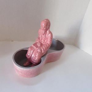 May include: A pink ceramic double dish with a figurine of a seated man with a beard and a hat. The figurine is in the center of the dish and is facing the left side of the image.