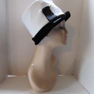 May include: A white hat with a black brim and a black ribbon, adorned with two small pearl-like embellishments. The hat is displayed on a mannequin head, showcasing its design and shape. The hat's design is a classic style.