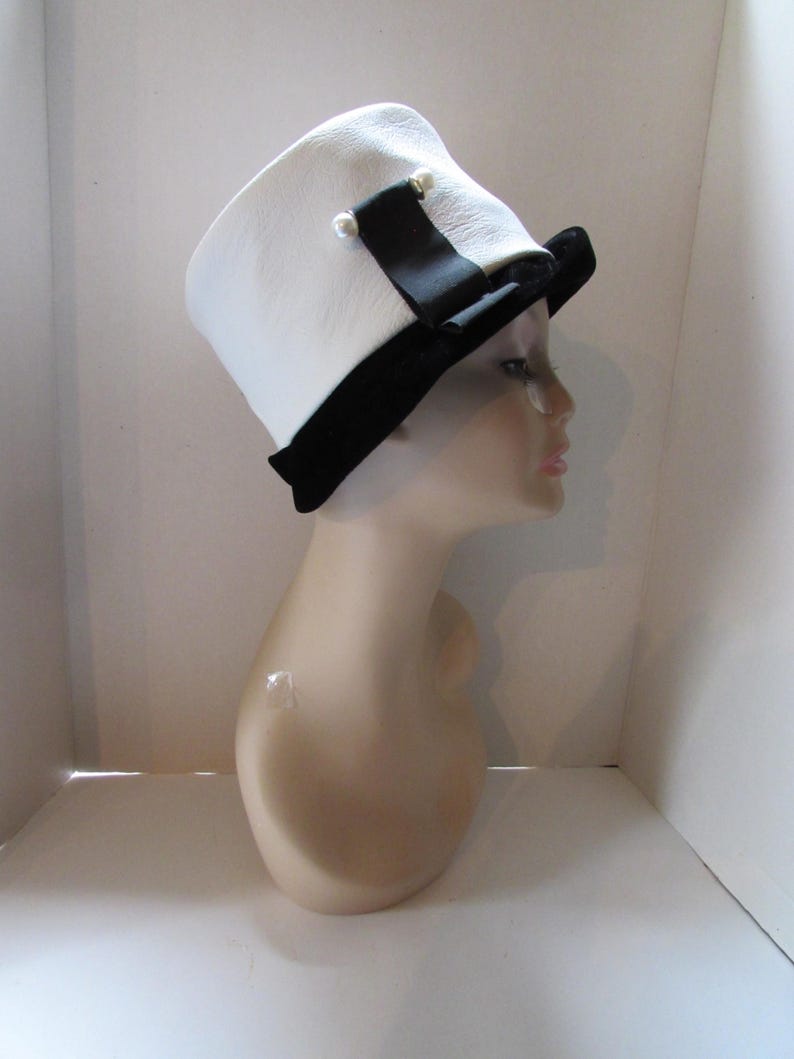 May include: A white hat with a black brim and a black ribbon, adorned with two small pearl-like embellishments. The hat is displayed on a mannequin head, showcasing its design and shape. The hat's design is a classic style.