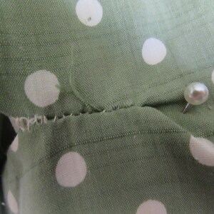 May include: Close-up of a green fabric with white polka dots. A white pearl-headed pin is inserted into the fabric.