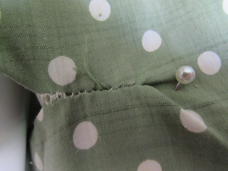 May include: Close-up of a green fabric with white polka dots. A white pearl-headed pin is inserted into the fabric.