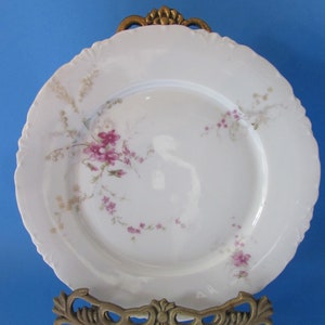May include: A white porcelain plate with a scalloped edge, adorned with pink floral patterns and gold accents. The plate is displayed on a decorative gold metal stand against a blue background. A vintage piece.