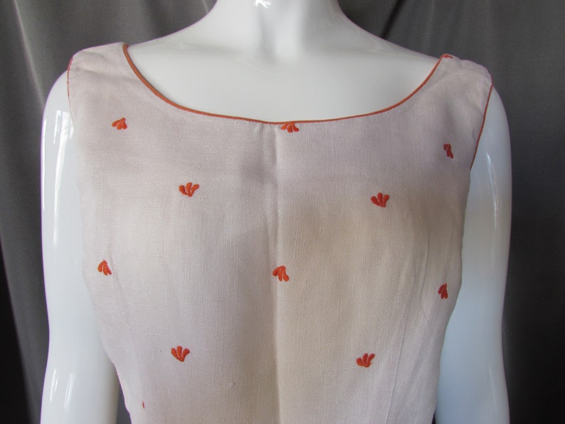 May include: A sleeveless white linen dress with a scoop neck and orange embroidered floral details.