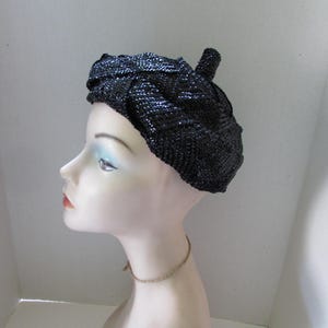 May include: A black beret featuring a textured, braided knit pattern with a subtle metallic sheen. The hat has a small, rounded pom-pom detail at the center top and is displayed on a mannequin head to show its rounded, classic shape and fit.