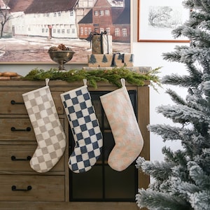 May include: Three checkered Christmas stockings hang on a wooden cabinet with a frosted artificial Christmas tree in the background. The stockings are beige and white, blue and white, and pink and white.
