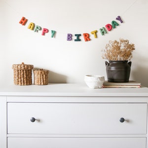 May include: A white dresser with two drawers and black knobs. The dresser is decorated with a felt banner that reads "Happy Birthday" in rainbow colors.