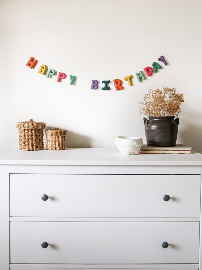 May include: A white dresser with two drawers and black knobs. The dresser is decorated with a felt banner that reads "Happy Birthday" in rainbow colors.