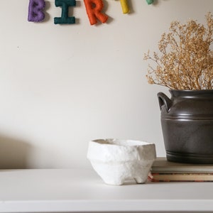 May include: A white paper mache bowl sits on a white dresser with a felt birthday banner hanging on the wall behind it. The banner reads "BIRTHDAY".
