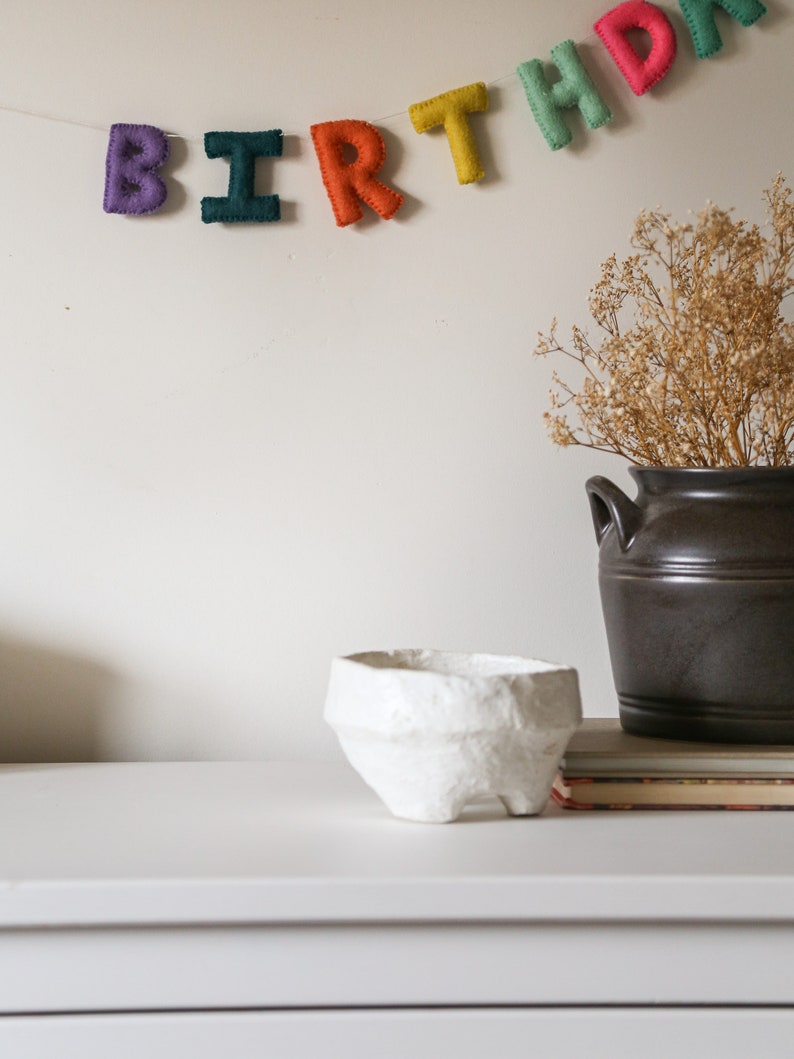 May include: A white paper mache bowl sits on a white dresser with a felt birthday banner hanging on the wall behind it. The banner reads "BIRTHDAY".