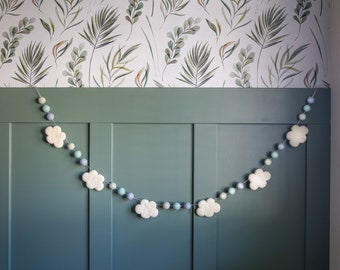 May include: A white felt cloud garland with blue and green felt balls hanging on a green wall with a white floral wallpaper.