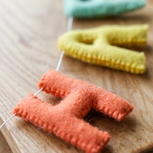 May include: A felt banner with the word "Happy" spelled out in colorful letters. The letters are strung together with white thread and are hanging on a wooden surface. The letters are in the colors pink, light blue, yellow, and orange.