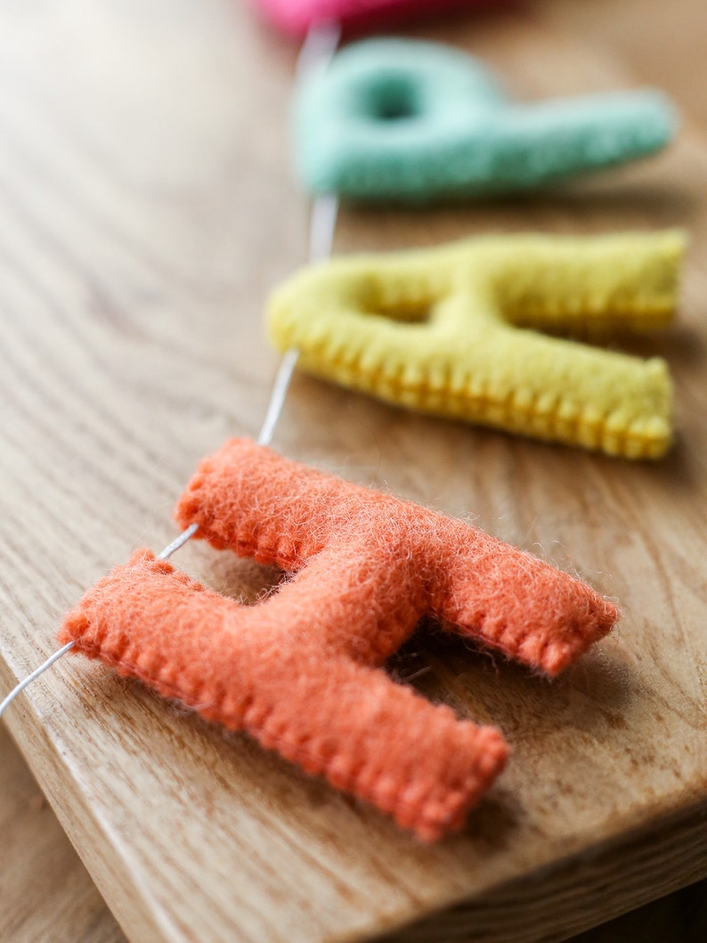 May include: A felt banner with the word "Happy" spelled out in colorful letters. The letters are strung together with white thread and are hanging on a wooden surface. The letters are in the colors pink, light blue, yellow, and orange.