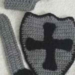 May include: A crocheted set consisting of a gray shield with a black cross emblem, a long gray sword with a black handle, and a gray helmet. Each piece is crafted from yarn with visible stitches, featuring a consistent gray and black color scheme.