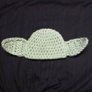 May include: A light green crocheted hat featuring a rounded crown and two large, pointed, protruding ears on either side. The item is crafted with a visible yarn stitch pattern, creating a textured appearance throughout the entire piece.