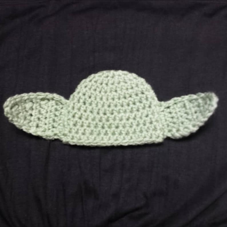 May include: A light green crocheted hat featuring a rounded crown and two large, pointed, protruding ears on either side. The item is crafted with a visible yarn stitch pattern, creating a textured appearance throughout the entire piece.