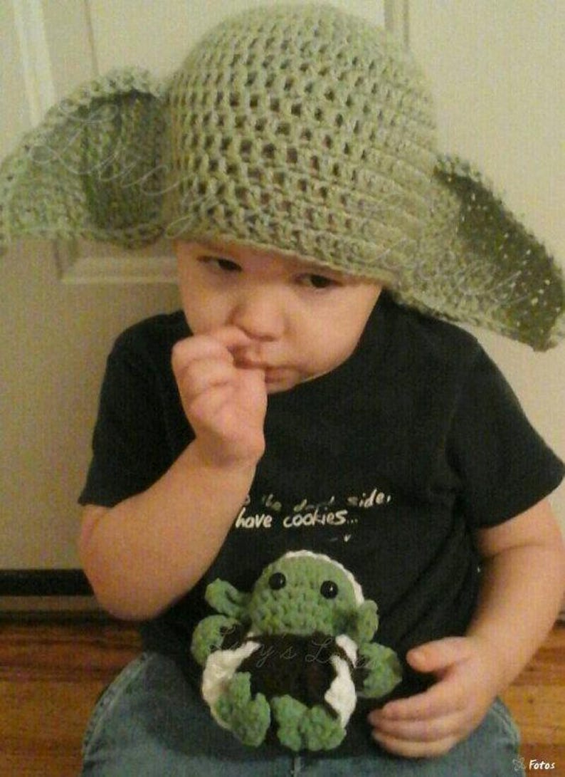 May include: A light green crocheted hat with large, pointed side flaps and a small, green and white crocheted creature toy. The hat features an open-weave stitch pattern, and the toy has dark eyes and a small body with green and white yarn details.