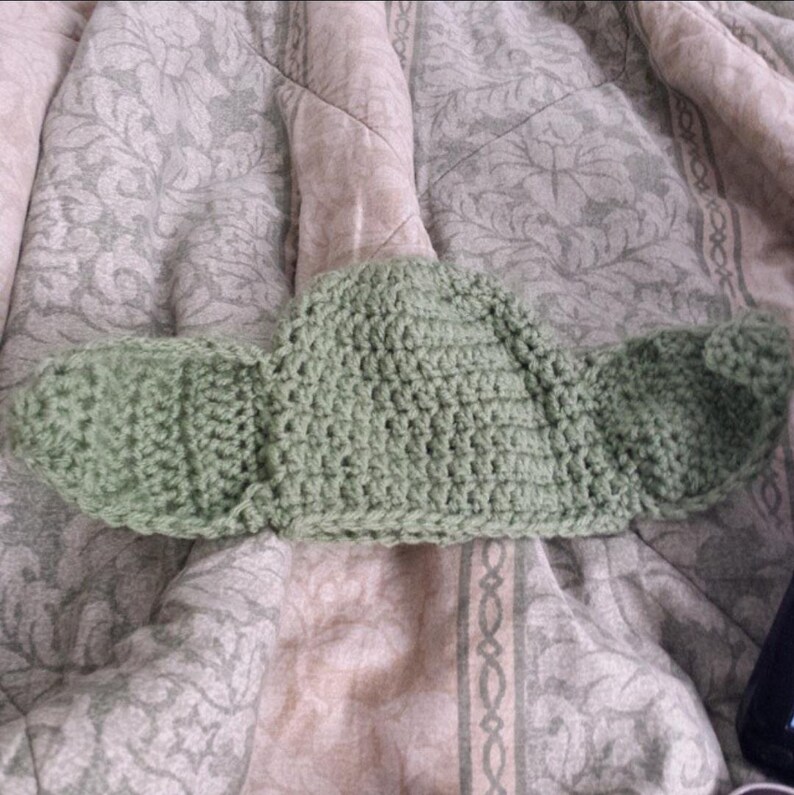 May include: A light sage green crochet hat featuring two large, rounded, protruding ear-like flaps on either side. The hat is constructed with a visible yarn stitch pattern, creating a textured surface throughout the entire piece.