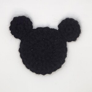 May include: A black crochet applique shaped like a circular head with two smaller circular ears attached to the top. The piece is crafted from yarn with a visible textured stitch pattern, creating a flat, silhouette-like design in solid black.