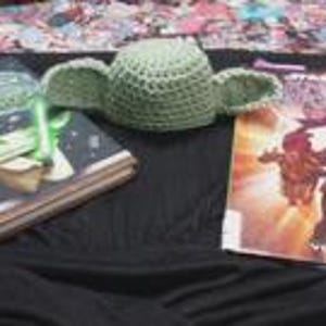 May include: A light green crocheted hat featuring two large, pointed, protruding ears. The hat is positioned between two books, one with a dark cover and a green light-up accessory, and another with a colorful illustrated cover featuring characters.