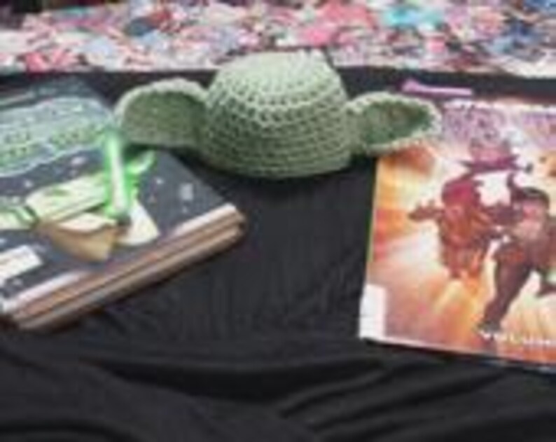 May include: A light green crocheted hat featuring two large, pointed, protruding ears. The hat is positioned between two books, one with a dark cover and a green light-up accessory, and another with a colorful illustrated cover featuring characters.