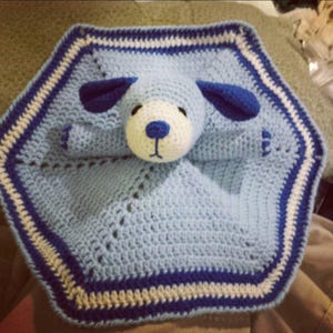 May include: A light blue and white crocheted security blanket featuring a central stuffed animal head resembling a dog. The blanket has a hexagonal shape with alternating stripes of light blue, white, and dark blue along the outer edges.