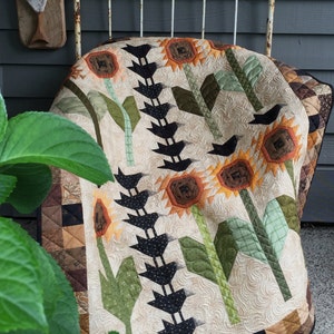 May include: A handmade quilt with a sunflower and bird design. The quilt has a beige background with appliqued sunflowers in shades of orange and brown, green leaves, and black bird accents. The quilt is draped over a vintage metal bed frame.