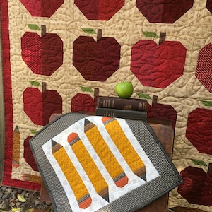 May include: A grey and white cushion with four yellow pencils on a white background. The cushion is sitting on a wooden bench in front of a red and green apple quilt.