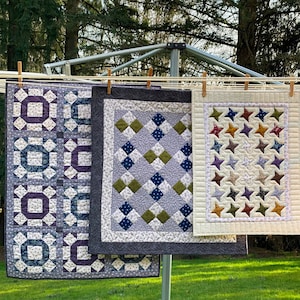 May include: Three handmade quilts hanging on a clothesline. The quilts are made with different fabrics and patterns. The first quilt is a purple and blue patchwork design. The second quilt is a gray and green diamond pattern. The third quilt is a cream colored quilt with a star pattern.