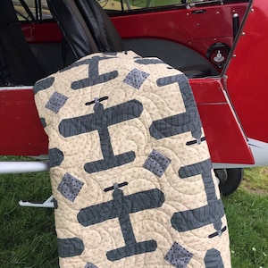 Puede incluir: Un edred&oacute;n beige y gris con tem&aacute;tica de avi&oacute;n. El edred&oacute;n presenta dise&ntilde;os de aviones grises repetidos y acentos cuadrados sobre un fondo beige. El edred&oacute;n est&aacute; colocado sobre el borde de un avi&oacute;n rojo.