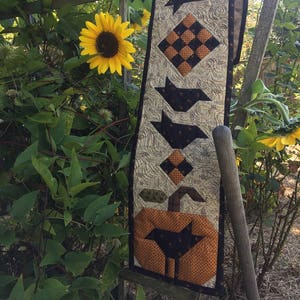 Könnte beinhalten: Ein schwarzer Vogel sitzt auf einem Holzzaun mit einem handgefertigten Halloween-Tischläufer mit einem Kürbis, schwarzen Vögeln und einem karierten Muster. Der Tischläufer ist aus Stoff und hat einen rustikalen, Vintage-Stil.