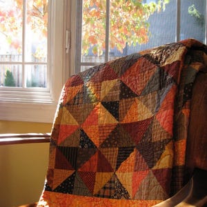 May include: A handmade quilt draped over a chair, featuring a patchwork design of triangles in warm autumn colors. The quilt's palette includes shades of orange, brown, and gold, creating a cozy and inviting aesthetic. The quilt is made of various patterned fabrics.