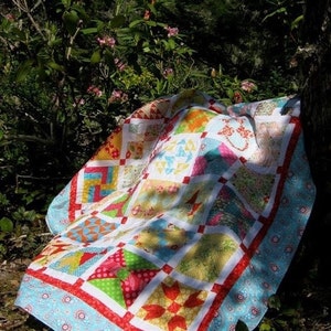 May include: A colorful patchwork quilt with a variety of floral and geometric patterns. The quilt is made with bright colors, including red, yellow, blue, green, and pink. It is laid out on the ground in a grassy area with a tree in the background.