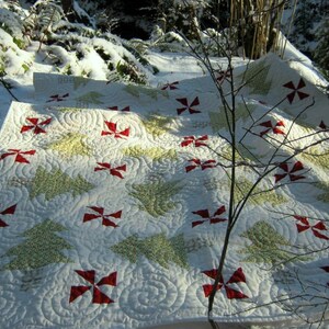 May include: A white quilt with red and white pinwheel and green Christmas tree designs. The quilt is placed on a snowy surface, with a background of snow-covered foliage and bare tree branches.