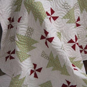 May include: A white quilt with a repeating pattern of green Christmas trees and red pinwheel shapes. The trees have a dotted pattern, and the quilt has a quilted texture. The quilt is folded, showing the design.