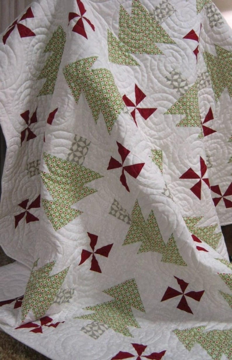 May include: A white quilt with a repeating pattern of green Christmas trees and red pinwheel shapes. The trees have a dotted pattern, and the quilt has a quilted texture. The quilt is folded, showing the design.