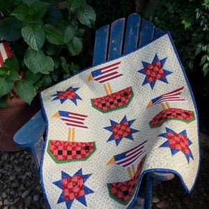 May include: A white quilt with a patriotic theme featuring red, white, and blue stars and watermelons. The quilt is folded and draped over a blue wooden chair.