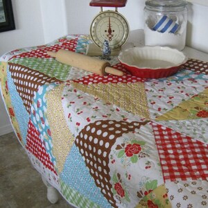 May include: A round, quilted tablecloth with a patchwork design. The fabric features various patterns, including polka dots, floral prints, and gingham checks in red, brown, yellow, and blue. A wooden rolling pin, a red pie dish, and a vintage scale are on the table.