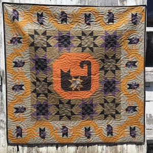May include: A quilted wall hanging with a black cat design in the center. The quilt is made with a variety of fabrics in shades of orange, brown, purple, and black. The quilt is surrounded by a border of orange and brown fabric.