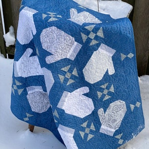 Op de afbeelding: Een blauwe en witte quilt met een patroon van wanten. De quilt is gevouwen en leunt tegen een houten hek.
