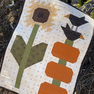 May include: A quilted wall hanging with a sunflower, a black bird, and three pumpkins. The background is a white fabric with a small black floral pattern.