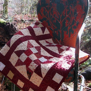 Puede incluir: Una colcha roja y blanca con un patrón floral está drapeada sobre una silla de metal vintage verde y roja. La silla está sentada en un área boscosa con hojas y ramas caídas.