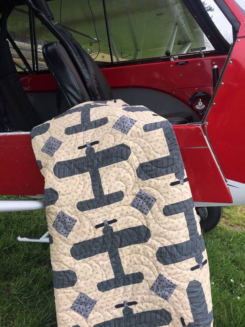 Puede incluir: Un edred&oacute;n beige y gris con tem&aacute;tica de avi&oacute;n. El edred&oacute;n presenta dise&ntilde;os de aviones grises repetidos y acentos cuadrados sobre un fondo beige. El edred&oacute;n est&aacute; colocado sobre el borde de un avi&oacute;n rojo.