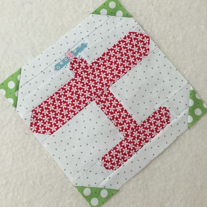 Puede incluir: Un bloque de patchwork hecho a mano con un estampado floral rojo sobre fondo blanco. El bloque presenta un dise&ntilde;o de avi&oacute;n estilizado con detalles en azul claro y rosa. Acentos de tela verde con lunares en las esquinas.