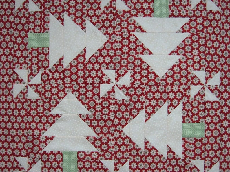 May include: A close-up of a quilt with a repeating pattern of white Christmas trees and pinwheels on a red background with small white and gray floral designs. The trees have green rectangular bases.