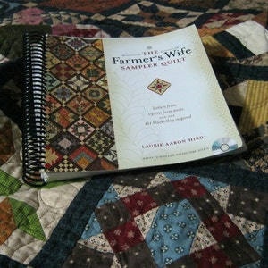 May include: A closed book titled "The Farmer's Wife Sampler Quilt" with a spiral binding. The book cover features a colorful quilt pattern and text that reads "Letters from 1920s farm wives and the 181 blocks they inspired." The book is lying on a patchwork quilt.