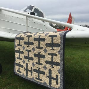 Puede incluir: Un edred&oacute;n hecho a mano con un patr&oacute;n repetido de aviones grises sobre un fondo crema. El edred&oacute;n est&aacute; colocado sobre el ala de un avi&oacute;n blanco en un campo de hierba. Tiene un borde azul oscuro.