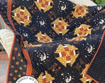 Puede incluir: Una colcha negra y naranja con un diseño de patchwork que presenta un patrón repetitivo de casas y estrellas. La colcha está hecha con una variedad de telas, incluyendo lunares, rayas y colores sólidos. La colcha está bordeada con un borde negro y naranja. La colcha está sentada en una silla de mimbre blanca. La colcha tiene una etiqueta que dice "Laugh Yourself Into Stitches".