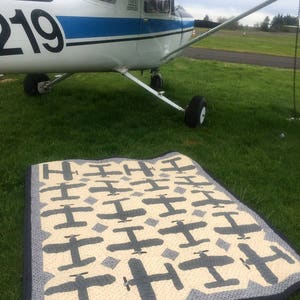 Puede incluir: Un avi&oacute;n blanco y azul est&aacute; estacionado en un campo cubierto de hierba. Una colcha hecha a mano con un patr&oacute;n de avi&oacute;n repetido en gris y crema est&aacute; extendida sobre la hierba frente al avi&oacute;n. La colcha tiene un borde azul oscuro.