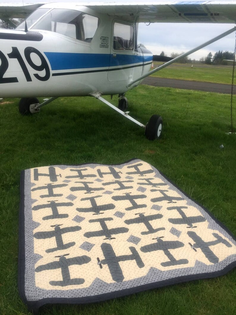 Puede incluir: Un avi&oacute;n blanco y azul est&aacute; estacionado en un campo cubierto de hierba. Una colcha hecha a mano con un patr&oacute;n de avi&oacute;n repetido en gris y crema est&aacute; extendida sobre la hierba frente al avi&oacute;n. La colcha tiene un borde azul oscuro.