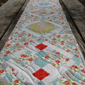 May include: A white table runner with a floral pattern in shades of red, blue, and green. The runner is quilted with a diamond pattern and has a scalloped edge.